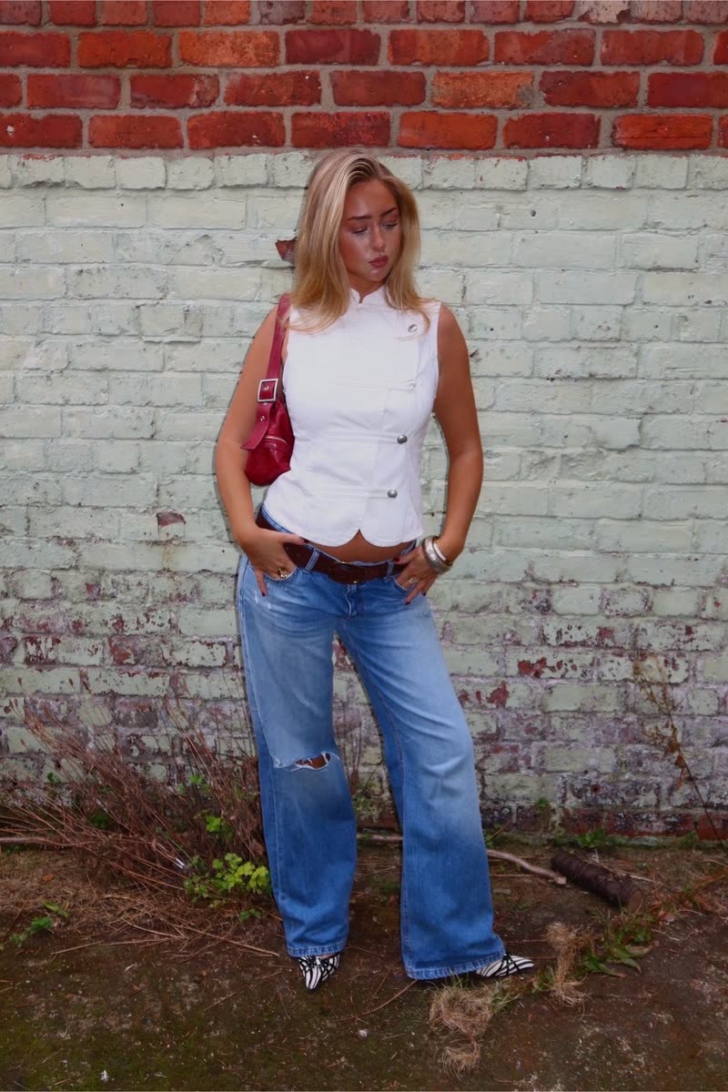 White waistcoat and wide-leg jeans with pointed shoes, against a painted brick wall